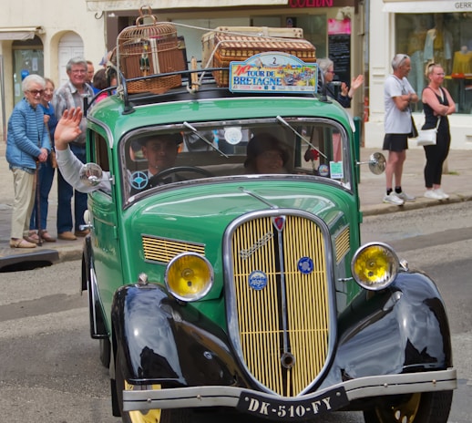 A vintage green car with a classic design is driving down a street. The car has luggage and birdcages on the roof, indicating a journey or road trip. People are gathered on the sidewalk, some waving, suggesting a festive or parade-like atmosphere. The sign on the car's roof reads 'Tour de Bretagne'.