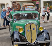 A vintage green car with a classic design is driving down a street. The car has luggage and birdcages on the roof, indicating a journey or road trip. People are gathered on the sidewalk, some waving, suggesting a festive or parade-like atmosphere. The sign on the car's roof reads 'Tour de Bretagne'.