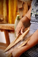 A person is using wooden tools to card fibers, suggesting a traditional textile process. The hands appear focused and engaged in the activity with strands of fibers visibly tangled between the tools. The background features blurred colors and shapes, with a hint of yellow yarn hanging.
