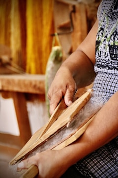A person is using wooden tools to card fibers, suggesting a traditional textile process. The hands appear focused and engaged in the activity with strands of fibers visibly tangled between the tools. The background features blurred colors and shapes, with a hint of yellow yarn hanging.