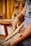 A person is using wooden tools to card fibers, suggesting a traditional textile process. The hands appear focused and engaged in the activity with strands of fibers visibly tangled between the tools. The background features blurred colors and shapes, with a hint of yellow yarn hanging.