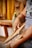 A person is using wooden tools to card fibers, suggesting a traditional textile process. The hands appear focused and engaged in the activity with strands of fibers visibly tangled between the tools. The background features blurred colors and shapes, with a hint of yellow yarn hanging.