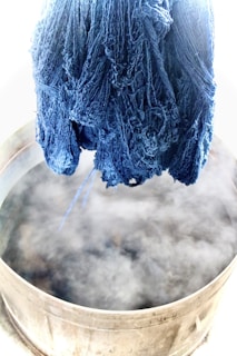 A bundle of blue yarn hangs suspended over a steaming metal vat. The scene suggests a dyeing process, with the yarn having a rich, deep indigo color. The steam is prominently rising from the vat, indicating heat and activity.