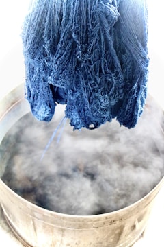 A bundle of blue yarn hangs suspended over a steaming metal vat. The scene suggests a dyeing process, with the yarn having a rich, deep indigo color. The steam is prominently rising from the vat, indicating heat and activity.