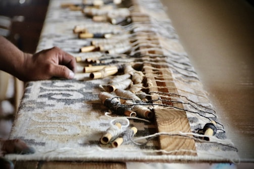 A hand is pictured reaching out to a large woven textile, with wooden and metallic spools scattered across it. The textile appears to be part of a traditional loom setup, indicating a crafting or weaving activity.