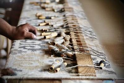 A hand is pictured reaching out to a large woven textile, with wooden and metallic spools scattered across it. The textile appears to be part of a traditional loom setup, indicating a crafting or weaving activity.