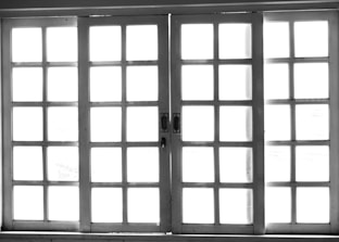 A set of white-framed sliding glass doors with multiple square panes, allowing bright light to pass through, creating a high contrast between the dark frame and the bright background.