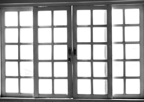 A set of white-framed sliding glass doors with multiple square panes, allowing bright light to pass through, creating a high contrast between the dark frame and the bright background.