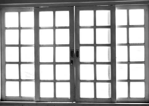 A set of white-framed sliding glass doors with multiple square panes, allowing bright light to pass through, creating a high contrast between the dark frame and the bright background.