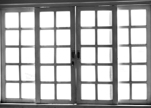 A set of white-framed sliding glass doors with multiple square panes, allowing bright light to pass through, creating a high contrast between the dark frame and the bright background.