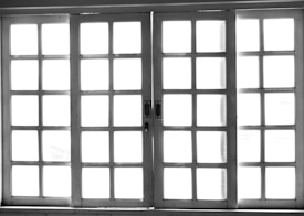 A set of white-framed sliding glass doors with multiple square panes, allowing bright light to pass through, creating a high contrast between the dark frame and the bright background.