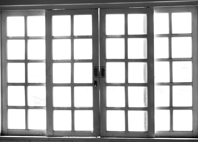 A set of white-framed sliding glass doors with multiple square panes, allowing bright light to pass through, creating a high contrast between the dark frame and the bright background.