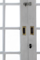 A close-up view of the central section of a white wooden double-door with multiple rectangular glass panes. The door features brass handles and a dark metal latch with a keyhole at the top.