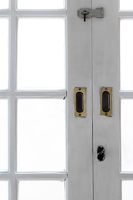 A close-up view of the central section of a white wooden double-door with multiple rectangular glass panes. The door features brass handles and a dark metal latch with a keyhole at the top.
