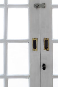 A close-up view of the central section of a white wooden double-door with multiple rectangular glass panes. The door features brass handles and a dark metal latch with a keyhole at the top.