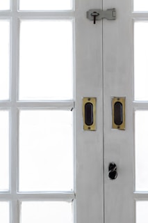 A close-up view of the central section of a white wooden double-door with multiple rectangular glass panes. The door features brass handles and a dark metal latch with a keyhole at the top.