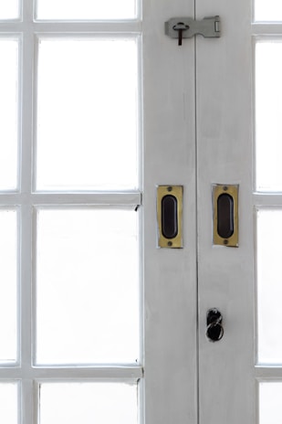 A close-up view of the central section of a white wooden double-door with multiple rectangular glass panes. The door features brass handles and a dark metal latch with a keyhole at the top.