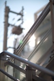 A close-up view of metallic railings and glass panels with sunlight creating reflections. A blurred background shows the outlines of an electrical pole and cables against a clear blue sky.