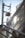 A close-up view of metallic railings and glass panels with sunlight creating reflections. A blurred background shows the outlines of an electrical pole and cables against a clear blue sky.