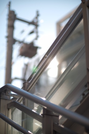 A close-up view of metallic railings and glass panels with sunlight creating reflections. A blurred background shows the outlines of an electrical pole and cables against a clear blue sky.