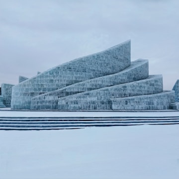 A large, modern architectural structure is constructed entirely from ice. It has multiple layered levels that curve upwards in a spiral-like design. The surrounding area is covered in snow, and the sky is overcast, giving a cold and serene atmosphere.