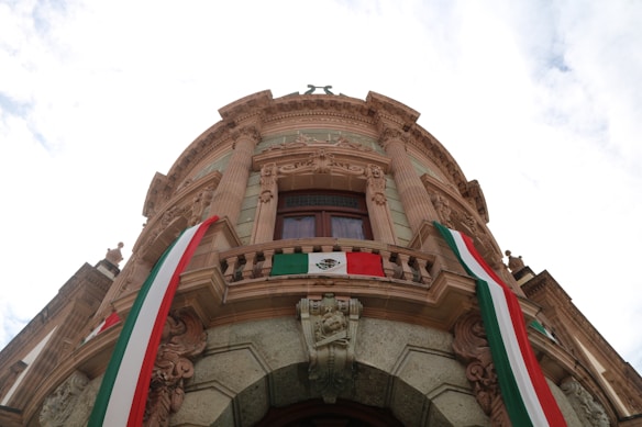A grand architectural building with ornate details and a central balcony. The structure is adorned with Mexican flags draped along the sides. The building features large columns and intricate carvings, with a prominent circular window on the upper level.