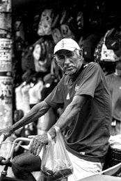 An elderly man wearing a cap and a sports jersey is seen holding a bicycle with one hand while clutching a plastic shopping bag with the other. He appears to be in a street market setting, surrounded by mannequins and various items like bags hanging in the background.