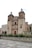 A large historic church with two prominent bell towers and detailed stonework stands at the center. In front of the church, there is a wide stone plaza bordered by agave plants. The sky is overcast, giving a soft light to the scene.