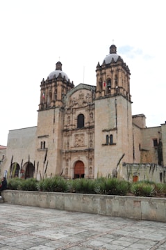 A large historic church with two prominent bell towers and detailed stonework stands at the center. In front of the church, there is a wide stone plaza bordered by agave plants. The sky is overcast, giving a soft light to the scene.