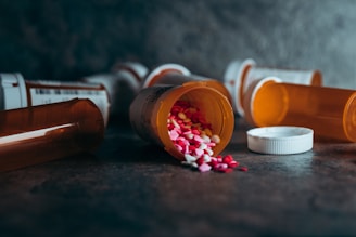 Several prescription pill bottles are lying on their sides on a dark surface, with one bottle in the foreground spilling out an assortment of colorful pills, including pink, yellow, and white. The scene is dimly lit, casting shadows around the bottles and creating a somber atmosphere.
