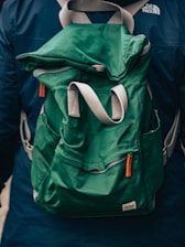 A green fabric backpack with adjustable gray and orange straps appears prominently. The backpack is worn by a person dressed in a dark blue jacket, evident from the partial view of shoulders. A logo is visible on the jacket.