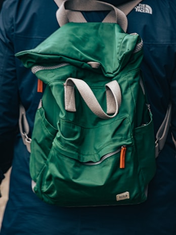 A green fabric backpack with adjustable gray and orange straps appears prominently. The backpack is worn by a person dressed in a dark blue jacket, evident from the partial view of shoulders. A logo is visible on the jacket.