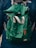A green fabric backpack with adjustable gray and orange straps appears prominently. The backpack is worn by a person dressed in a dark blue jacket, evident from the partial view of shoulders. A logo is visible on the jacket.
