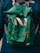 A green fabric backpack with adjustable gray and orange straps appears prominently. The backpack is worn by a person dressed in a dark blue jacket, evident from the partial view of shoulders. A logo is visible on the jacket.