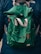 A green fabric backpack with adjustable gray and orange straps appears prominently. The backpack is worn by a person dressed in a dark blue jacket, evident from the partial view of shoulders. A logo is visible on the jacket.
