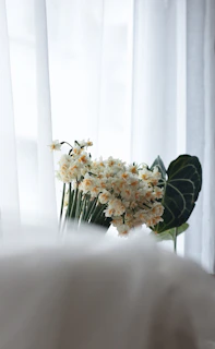 A serene morning light filtering through creamy curtains over a softly arranged bouquet of wildflowers on a wooden table.