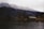A serene lakeside scene featuring several rustic wooden cabins nestled at the edge of calm water, set against a backdrop of snow-dusted, forested mountains. The overcast sky adds a moody atmosphere to the scene, while bare trees indicate a late autumn or early winter season.