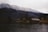 A serene lakeside scene featuring several rustic wooden cabins nestled at the edge of calm water, set against a backdrop of snow-dusted, forested mountains. The overcast sky adds a moody atmosphere to the scene, while bare trees indicate a late autumn or early winter season.