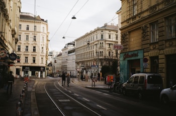 An urban street scene in a European city featuring historical buildings with ornate architecture. The street is lined with shops and cafes, with several people walking and bikes parked along the sidewalk. Tram tracks run down the center of the street.