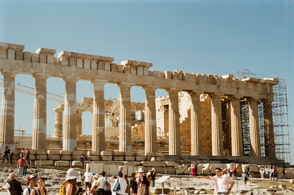 An ancient Greek temple with tall columns dominates the scene, showcasing classical architecture under a clear blue sky. Numerous tourists are walking around the structure, which appears to be partially under restoration, as indicated by scaffolding.