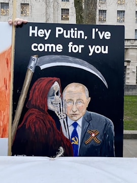A painted image features a figure in a red cloak with a skeletal face, representing the Grim Reaper, holding a scythe and standing next to a man in a suit with a somber expression. Text above reads, 'Hey Putin, I've come for you.' The man wears a suit with a tie displaying a Soviet hammer and sickle symbol, while an orange and black ribbon is attached to his suit.