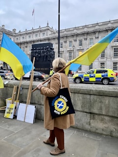 A person stands holding a large Ukrainian flag in front of a historic building. Nearby, other flags and placards are leaning against a wall. A black statue commemorating women of World War II is visible. In the background, police vehicles and an unseen monument can also be seen.