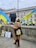A person stands holding a large Ukrainian flag in front of a historic building. Nearby, other flags and placards are leaning against a wall. A black statue commemorating women of World War II is visible. In the background, police vehicles and an unseen monument can also be seen.