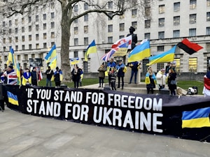 A group of people are gathered in a public square, holding various flags including the Ukrainian national flag and a British flag. They are standing behind a large black banner that reads, 'IF YOU STAND FOR FREEDOM STAND FOR UKRAINE'. The background features a large building with multiple windows and a statue of a soldier.