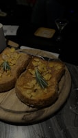 Two slices of toast topped with sliced almonds and garnished with rosemary, placed on a round wooden board. The setting appears to be a dimly lit dining table with a plate and glassware in the background.
