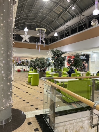 An interior view featuring a shopping mall or indoor public space with a high ceiling and large star and ornament decorations. Green chairs and tables are arranged in the seating area, and a few people are seated in the background. The space is well-lit with overhead lights and decorative string lights.