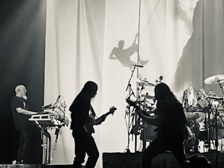 A black and white image of a live music performance featuring several musicians. One individual plays the keyboard while two others are seen playing guitars, surrounded by drums. There's a prominent shadow of a person in a climbing pose projected on a curtain in the background.