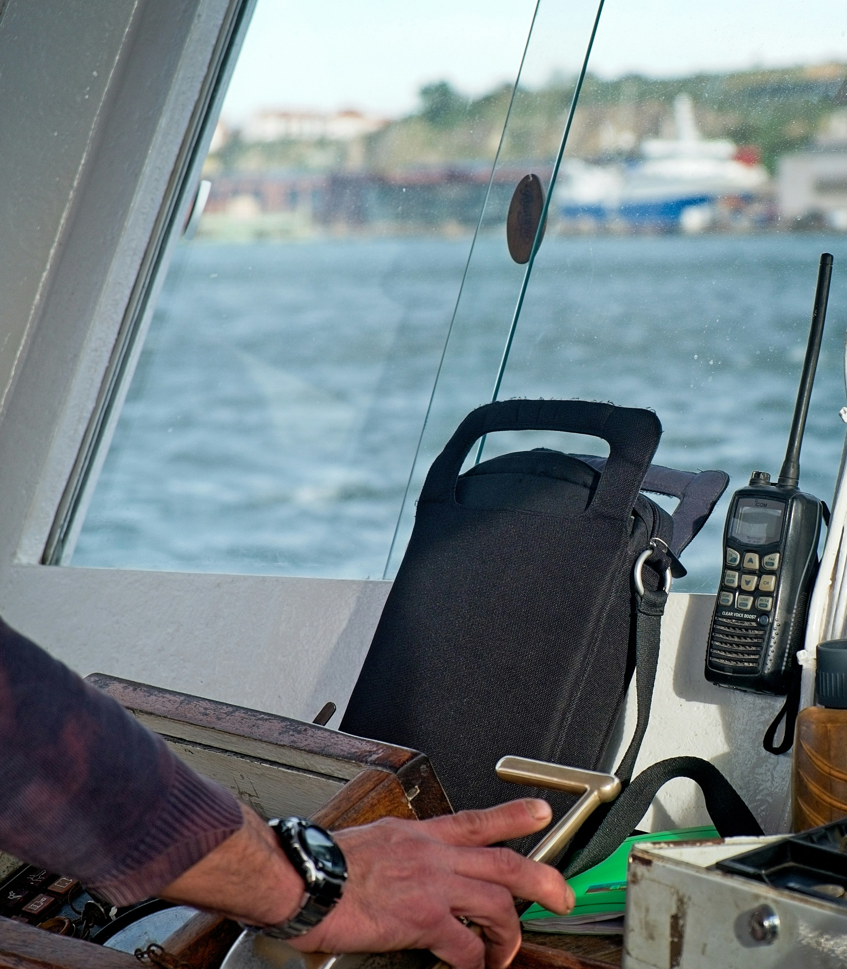 Black handheld marine radio with a long antenna propped at the windshield of a boat's console beside a dark gear bag, blue water outside