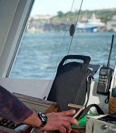 Black handheld marine radio with a long antenna propped at the windshield of a boat's console beside a dark gear bag, blue water outside