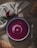 A bowl of vibrant beetroot soup is placed on a rustic wooden table. The soup has a deep magenta color with a swirl of cream on top. Surrounding the bowl are a whole beetroot, a few pieces of bread, a green leaf, and a silver spoon. A beige cloth is casually draped, adding to the warm, homely feel.